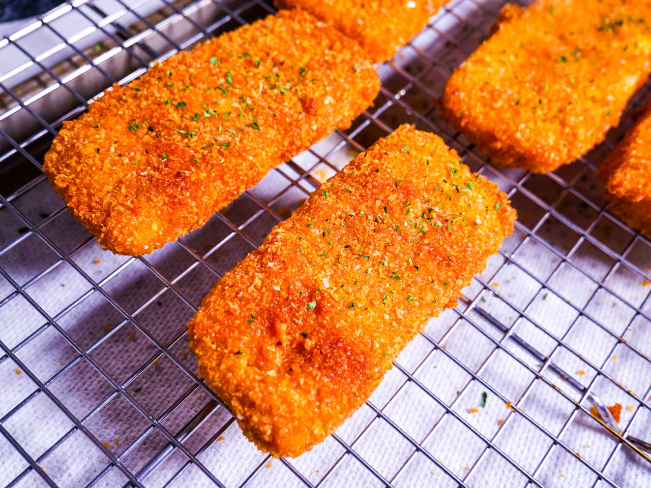 Fried mozzarella sticks cooling on a wire rack with paper towel lined baking sheet below.