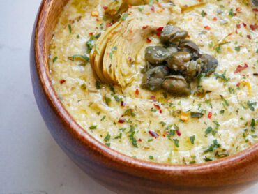 Creamy Artichoke Sauce with Capers in a wooden bowl on a white countertop with tarragon and red chili pepper garnish.