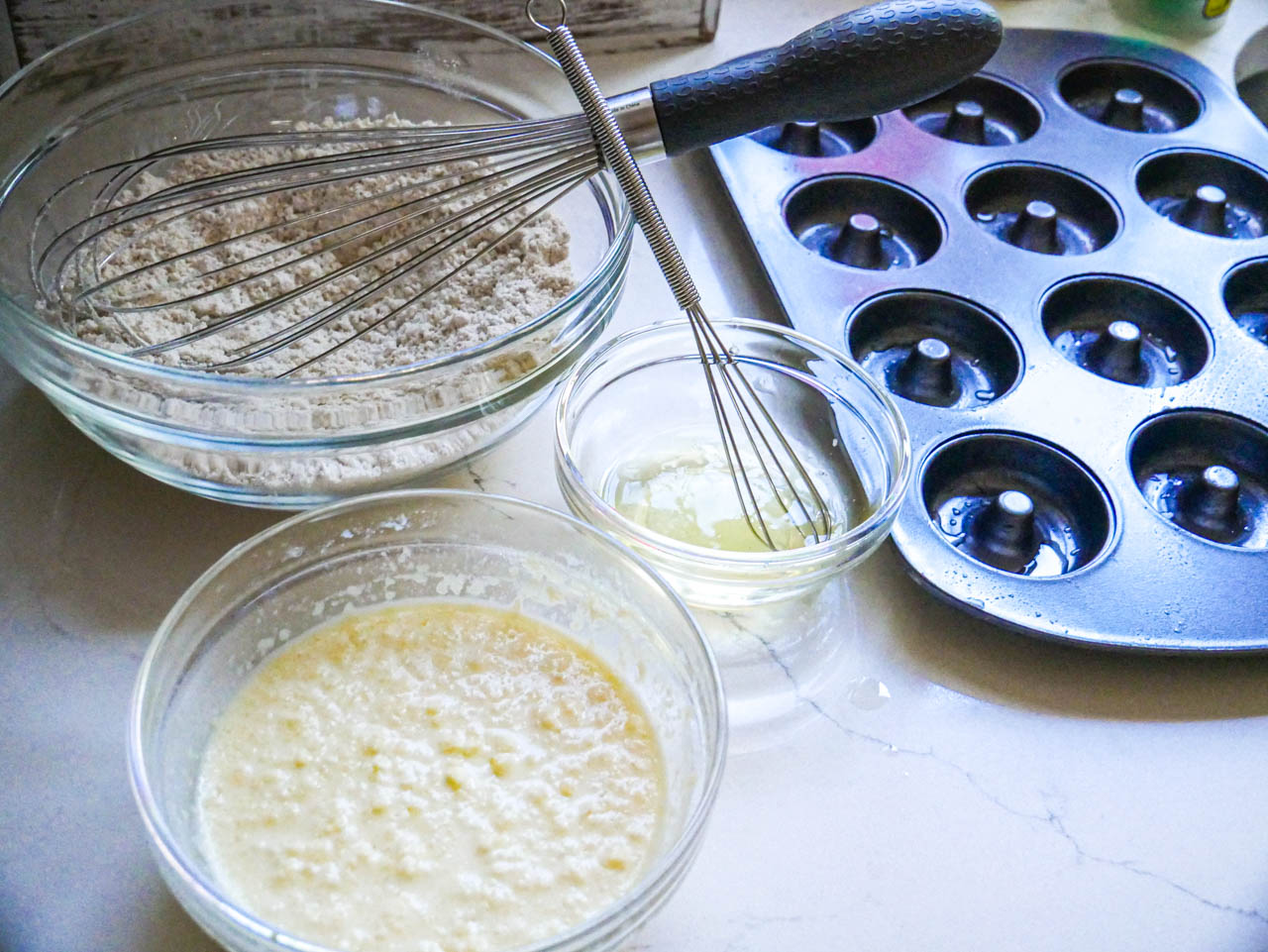 Flour and whisk in a bowl, wet ingredients in another bowl, egg whites in a small bowl on a white counter top next to a mini donut pan.