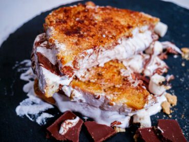 S'mores French Toast cut in half on a black circular stone plate with chocolate and fluff spread