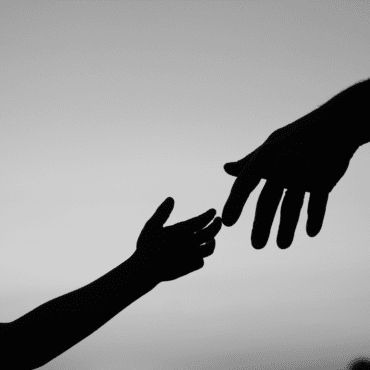 Grayscale photo with black silhouettes of adult hand and child hand reaching towards each other against a gray background.