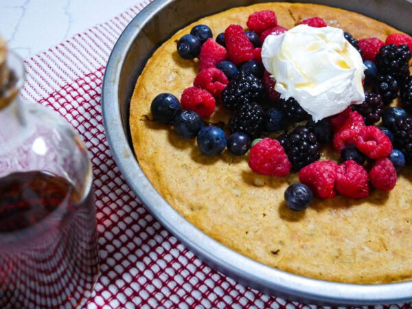 Tiger nut flour oven cake topped with berries and whipped cream in a cake pan, on a red and white placement with maple syrup in a bottle on the side.