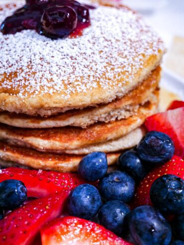 Tiger Nut Flour Pancakes. Blueberries and strawberries around the base and powdered sugar and blueberry sauce on a stack pancakes on a white plate.