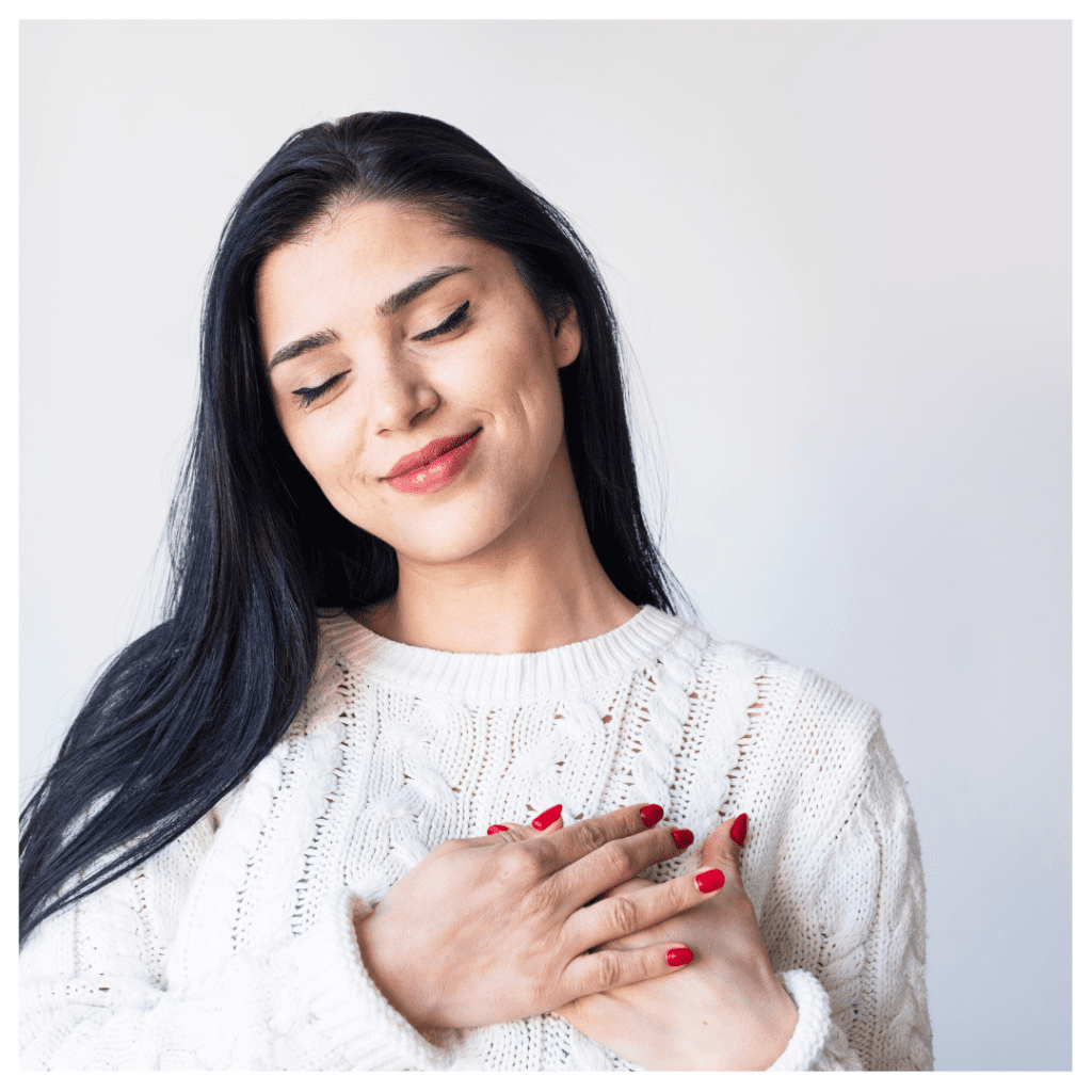 Self-respect in a relationship - woman in a white sweater, holding her hands to her heart against a white background.