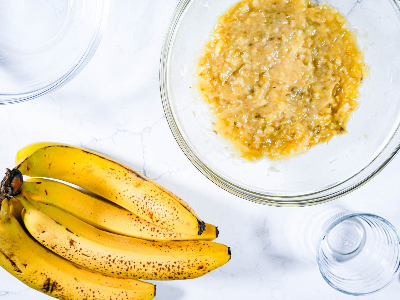 Mashed bananas in a clear bowl on a white countertop surrounded by other clear bowls and a ripe banana bunch.