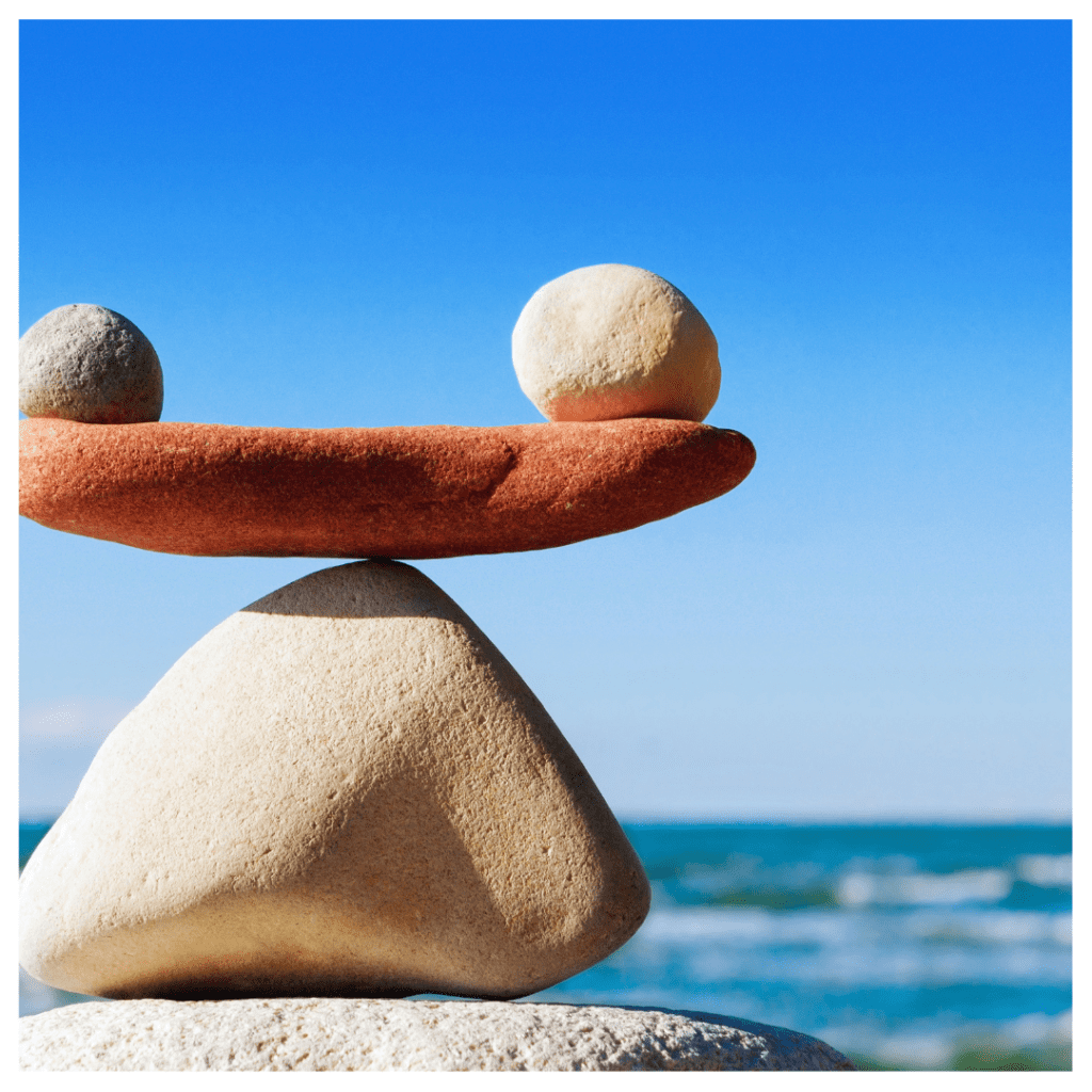 Two rocks balancing on one slab of red rock that's also balancing on a triangular shaped rock against an ocean background. Finding harmony with masculine and feminine energy.