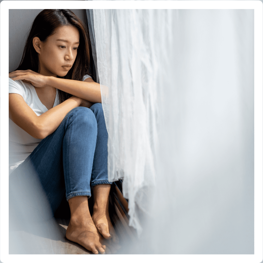 Woman in a white shirt and jeans in a room with natural light, hugging her shoulders. She looks sad as she sits next to shredded white curtains. She looks like she has emotional numbness.