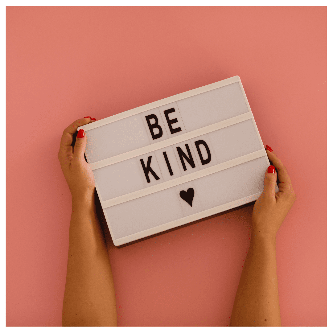 Pink background with a a sign that's held by a pair of hands. The sign says "Be Kind" with a heart. Kindness is encouraged.