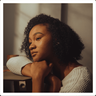 Woman sitting and leaning on table looking out the window in a beige outfit. She's in a dimly lit room displaying emotional numbness.