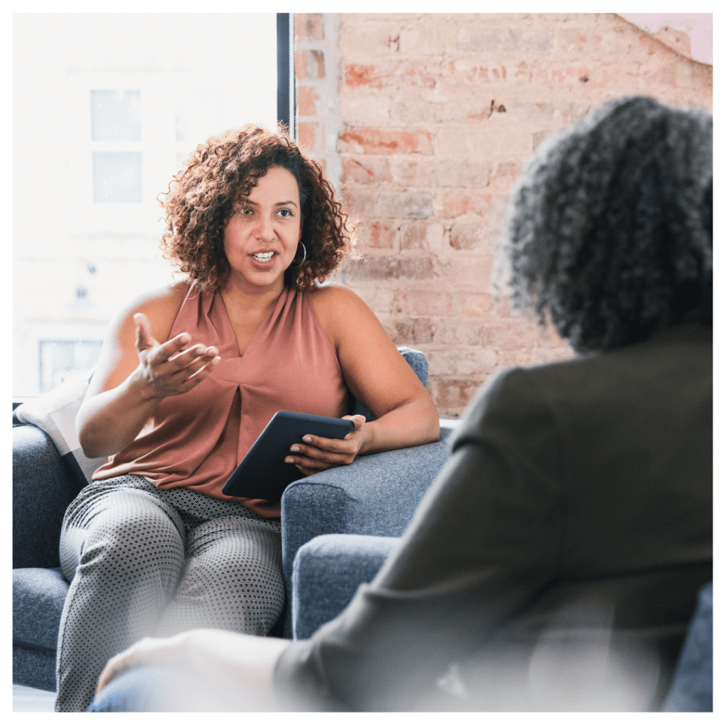 Two women are sitting in armchair, one woman advising the the other and facing the camera while the other has her back to it. 