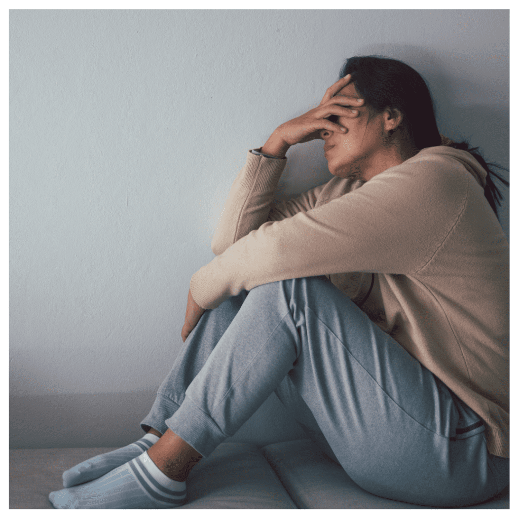 A woman overwhelmed with anxiety, hugging her knees as she sits on the couch. She's covering her eyes with one hand.