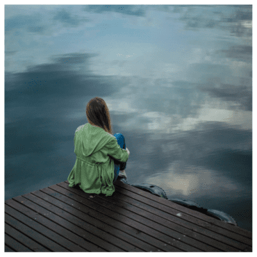 A girl sitting at the edge of a dock with water reflecting the dark clouds from the sky.