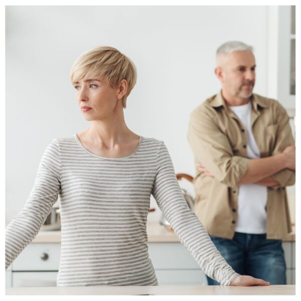 A woman looking out the window while her spouse is behind her with crossed arms signaling dismissive communication between the two